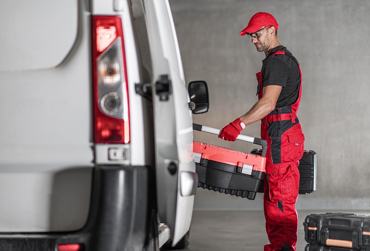 Man loading tool boxes into a van wearing red overalls, gloves and baseball cap