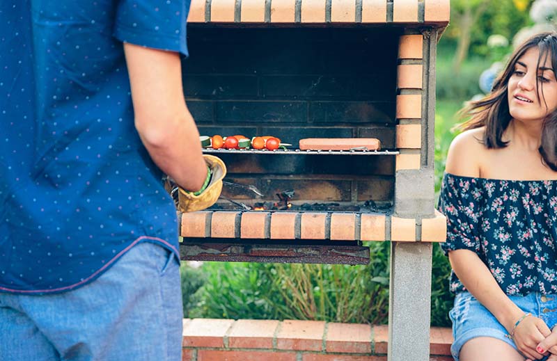 Person cooking sausages and vegetable kebabs on a brick BBQ with a lady sat to the side watching